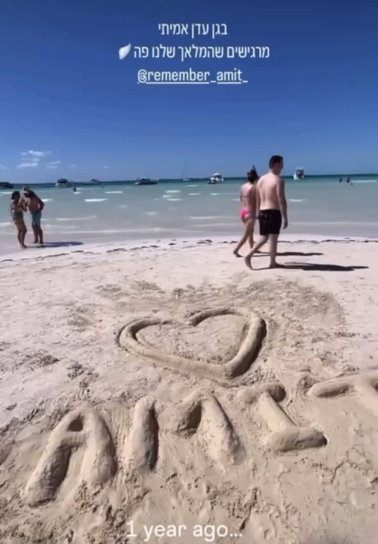 השם ״עמית״ כתוב באותיות באנגלית בחול על חוף הים, מים תכולים ברקע