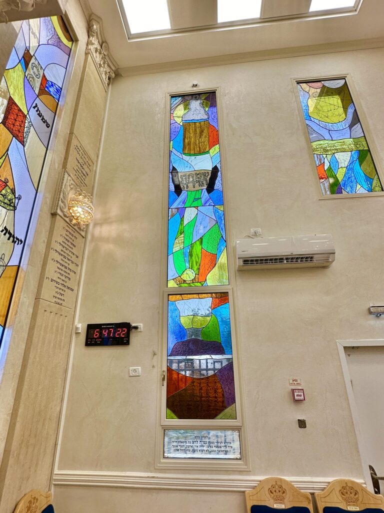 חלון ויטראז׳ צבעוני בתוך מבנה, לצידו לוח הקדשה